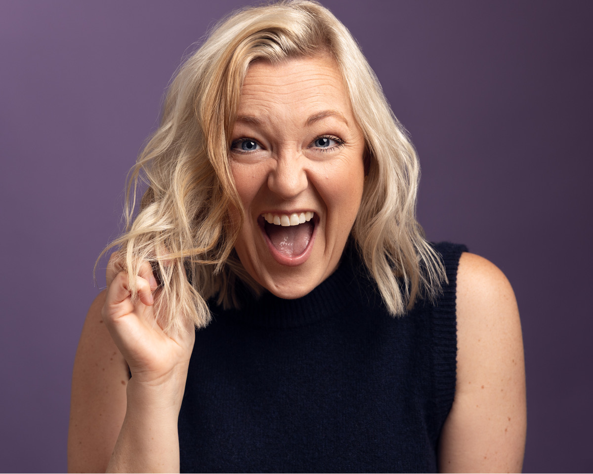A headshot of a person wearing a black top. They have shoulder length blonde hair. They pull an excited face and have one hand raised towards their face. The shot is taken against a purple background.