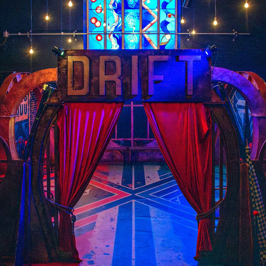 A sign saying drift above some red curtains. The curtains draw you into a space behind lit with a stain glass window.