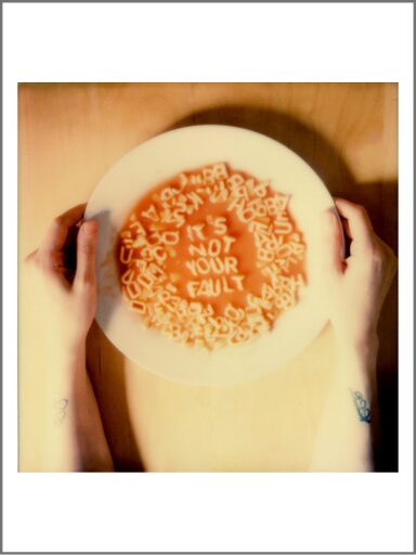 A photograph. Viewed from above, two hands gently hold a white pasta bowl filled with alphabet soup. The image has a warm, soft-focus quality with golden lighting. Scattered pasta letters float in the reddish-orange tomato sauce, but in the middle the letters are carefully arranged to form the message: “IT’S NOT YOUR FAULT.” The bowl sits against a pale wooden surface of a table, and faint tattoos are visible near both wrists.