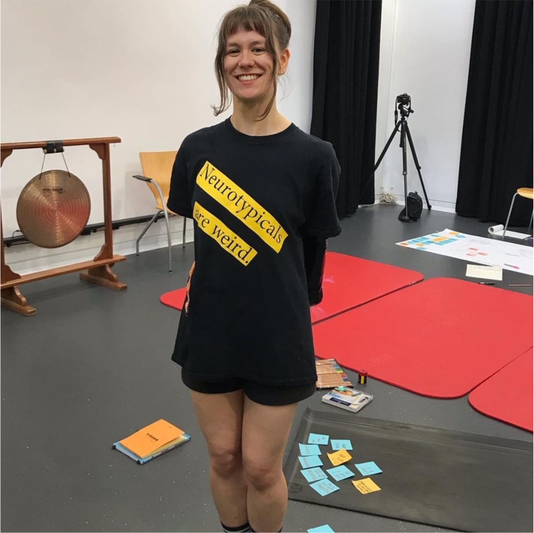 Aby wearing black shorts and tshirt with slogan 'Neurotypicals are weird'. Red yoga mats and gong in background