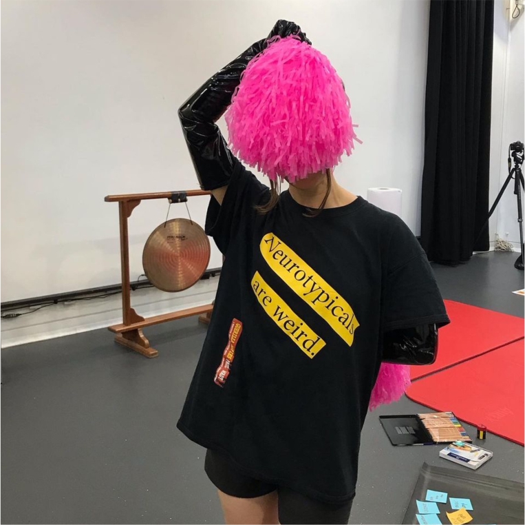 Aby Watson holding pink pom pom over their face and wearing black shorts and tshirt with slogan 'Neurotypicals are weird'. Large gong and red yoga mats on the floor in the background.