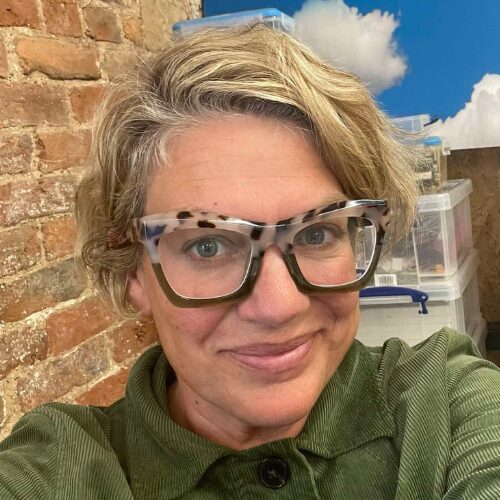 A headshot of Abbie Canning. They have short dark blonde hair. They wear thick rimmed glasses and a green top. They stand against a brick wall.