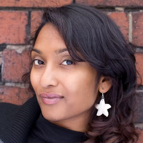 A headshot of Debbie Bandara. Debbie has dark shoulder length hair. They look sideways at the camera. They wear a white star shaped hanging ear ring and a black top and jacket. They stand against a brick wall.