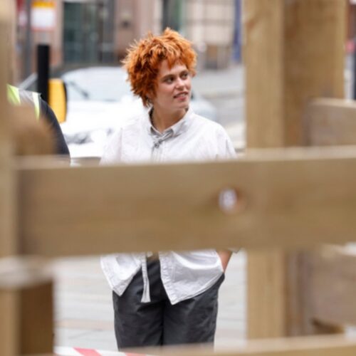 Maya Edwards standing in an urban location. They have short orange. They wear a white shirt and black trousers, with their hand in their pockets. In the foreground is an out of focus wooden structure.