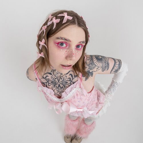 An image of Kao Hove aka kaoti standing against a white background. They have light brown shoulder length hair with pink bows in it, and wear a pale pink strappy top and fluffy pink leg warmers. They have dramatic dark pink eye make up and red and white freckles drawn on their face.