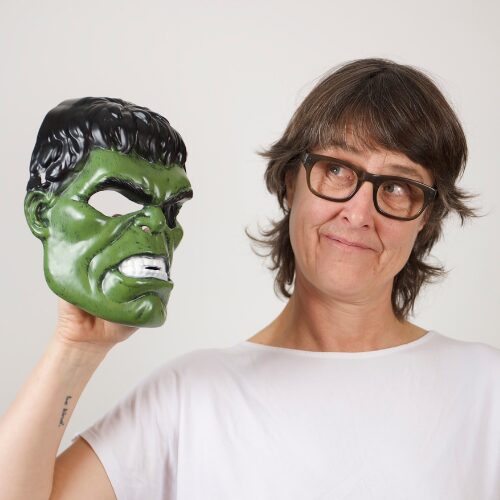 A headshot of Imogen Ashby. Imogen holds a hulk mask in her right hand. She has short dark tousled hair and wears glasses and a light coloured t shirt.