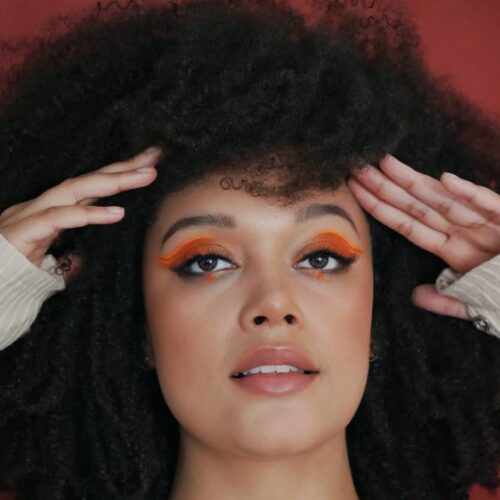 A headshot of Elle Chante, a mixed raced woman in her mid twenties with a large afro, against a terracotta backdrop. Her makeup is dramatic, with orange eyeshadow and orange rhinestones just below her eyes. She touches her forehead with her hands. Credit: Elle Chante Photography