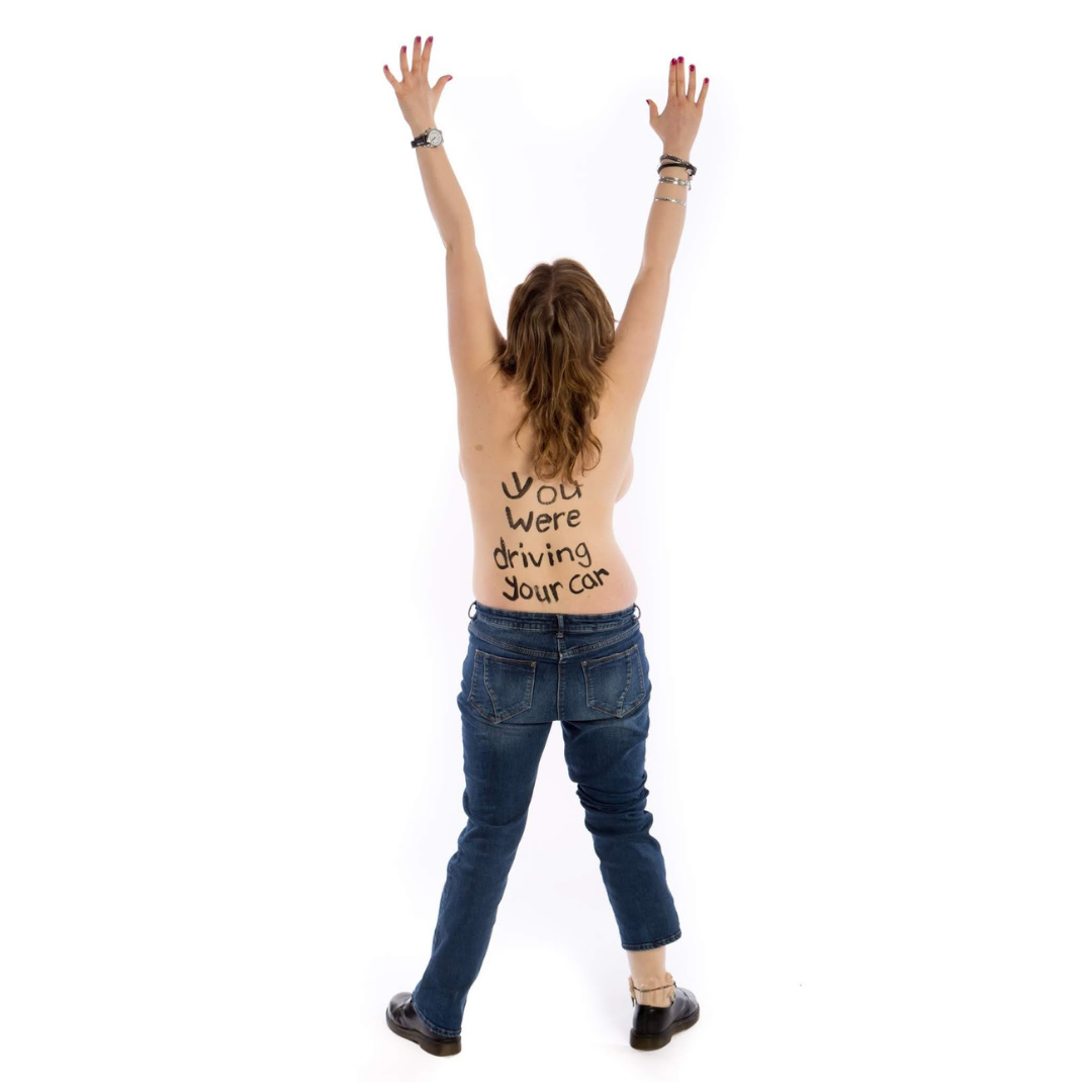 Grace Currie 1080x1080 A person with their arms upstretched facing away from the camera. They are wearing dark blue jeans. Written on their back in black pen is the statement - You were driving your car.