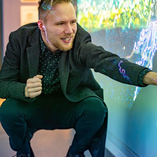 Henry Driver kneels down and gestures toward a large interactive screen showing colourful, wave-like graphics, while smiling in a bright indoor space.