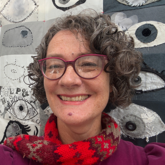A headshot of Lois Blackburn. Lois stands against a black and white quilt. Lois has short curly hair and wears red glasses, a red colourful scarf and a burgundy top.