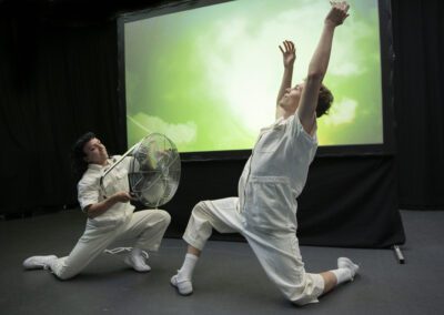 In a dark room two people in white boiler suits perform a dance routine. They are both crouched low, one extends their arms upwards while the other directs a portable fan towards them. Behind them a projector screen shows clouds in the sky.