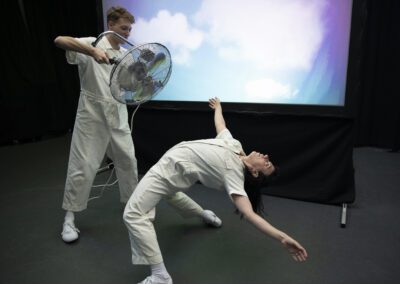 In a dark room two people in white boiler suits perform a dance routine. One is bent backwards with arms extended and staring at the ceiling while the other stands and points a portable fan towards them. A projection screen in the background displays clouds in the sky.