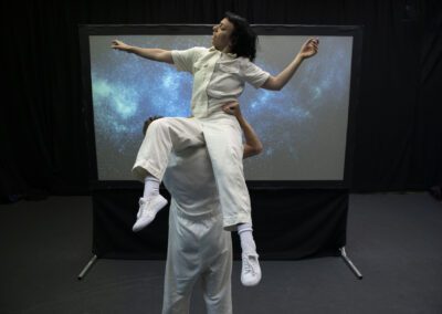 In a dark room surrounded by black curtains a central screen displays a projection of the night sky. In front of the screen are two people in white boiler suits, one stands facing the screen and lifting the other unto their shoulders.