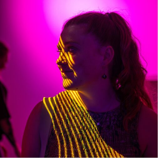 Hannah Fowkes in profile with a ponytail, lit by intense magenta and yellow light, wearing a dark top and earrings, with a yellow patterned overlay across the face and chest.