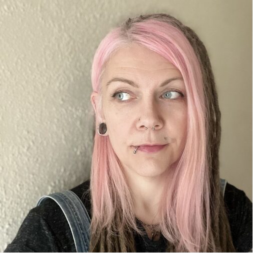 Headshot of Helen Jackson with long pastel pink hair, blue eyes, and facial piercings, wearing a black top and small earrings, against a light textured wall.