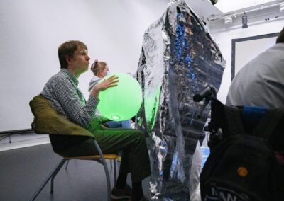 A person sits in a white room holding a bright green illuminated inflatable ball. People are seated either side of them and directly in front of them is a standing figure completely covered in a sheet of reflective metallic material.