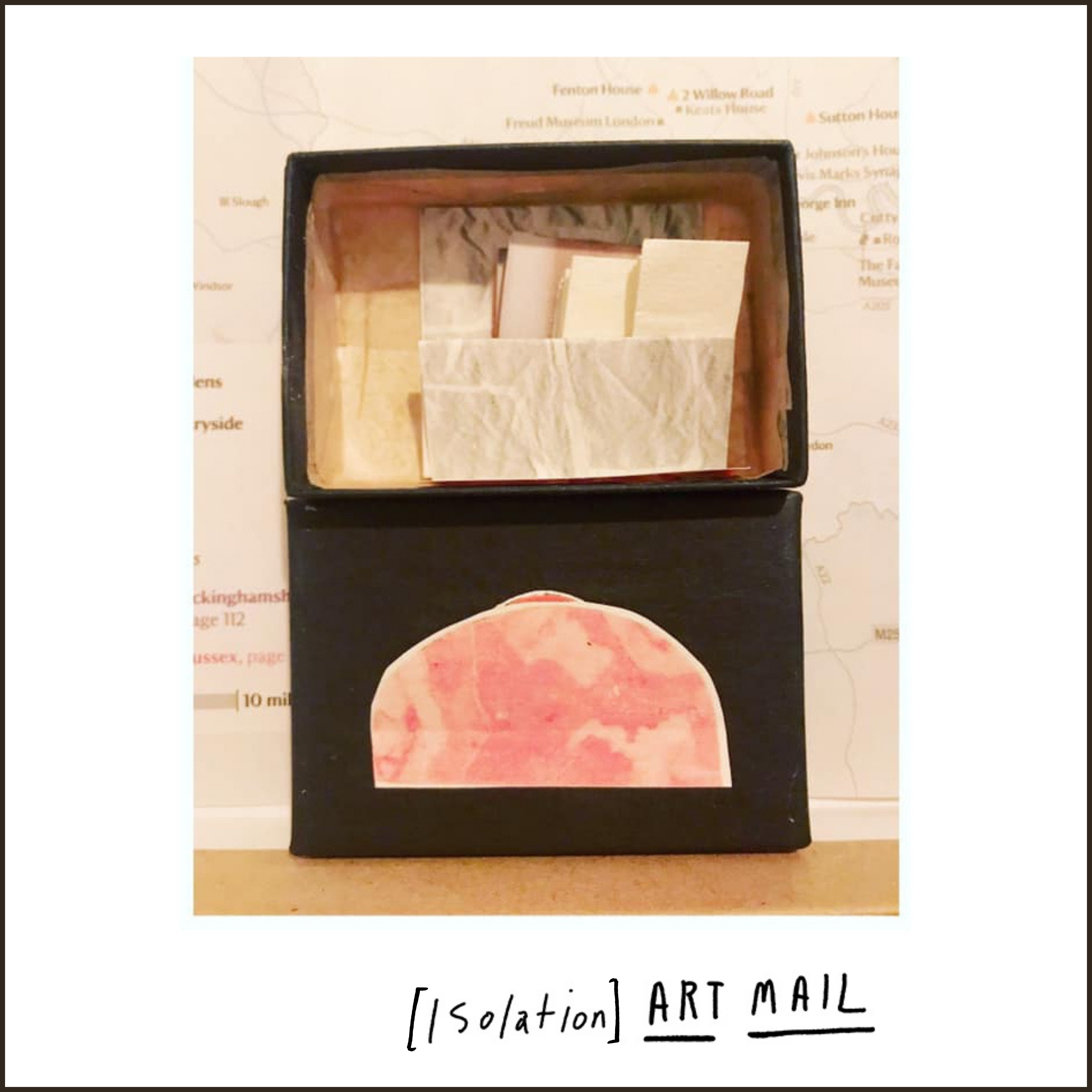 Lead image for an exhibition. A white background with text that reads [Isolation] Art Mail is handwritten in black at the bottom of the screen. In the centre, what looks like a black lunchbox stands upright against a cream map. Inside the box are paper versions of cheese and ham.
