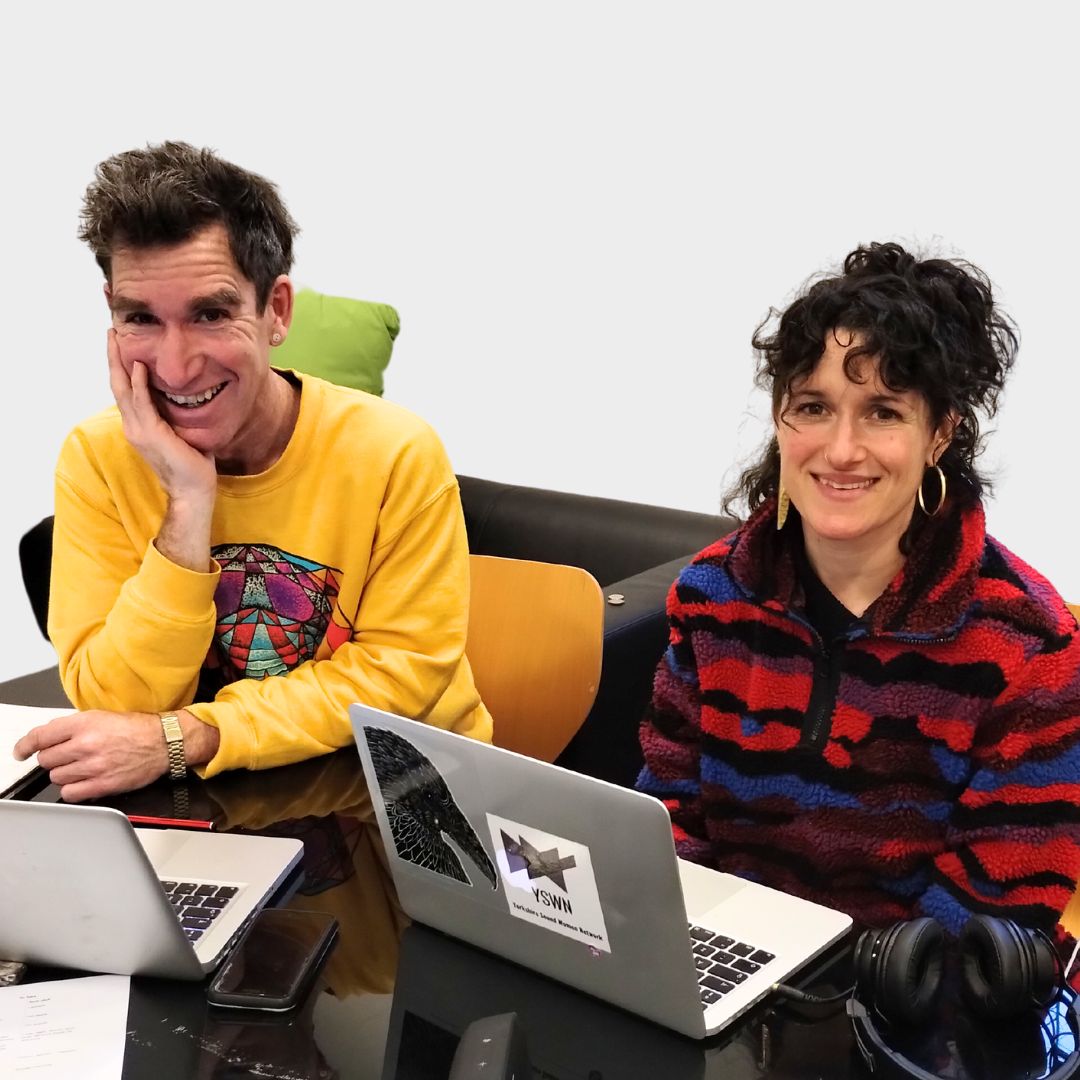 Two people sit at a desk smiling at the camera. They are working on laptops.