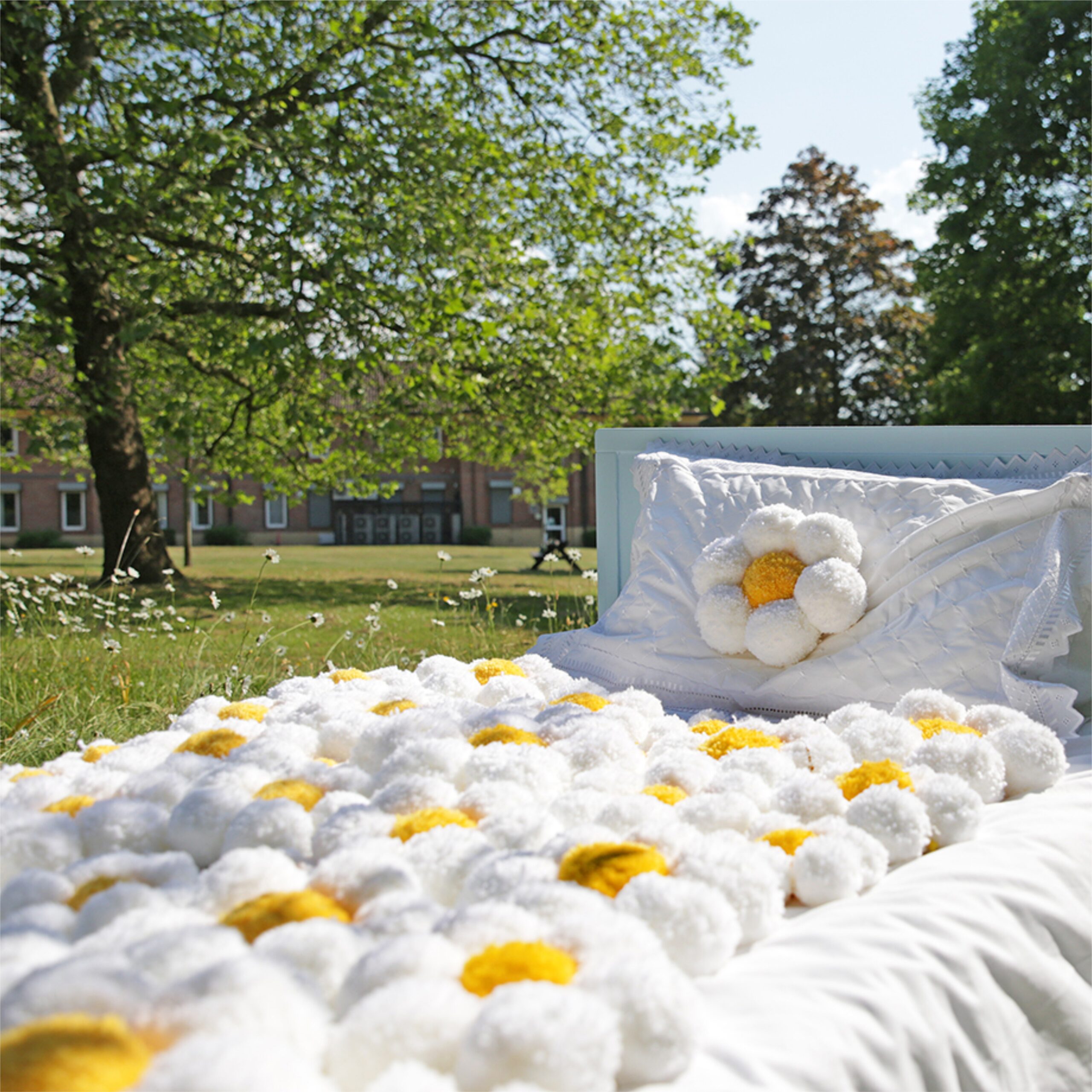 Bed outside in a field. On the bed is white pillow and quilt covered in white and yellow pom poms in the shape of daisies. In the background is grass and trees.