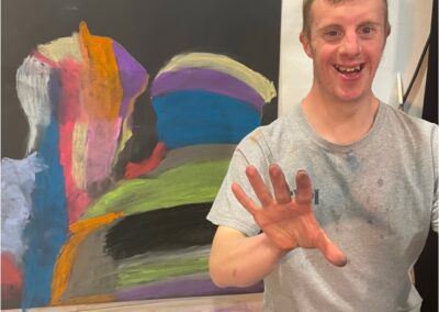 artist smiling at the camera wearing grey tshirt and holding up one hand covered in coloured chalk. Chalk sihouettes on black background on wall behind artist.
