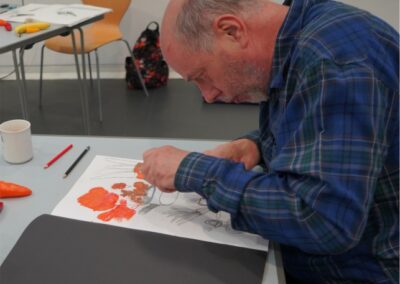 Older with grey hair artist wearing blue checked shirt drawing still life image with orange crayons