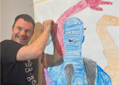 Artist wearing black tshirt with white writing, smiling at the camera and tracing shadow on the wall in blue, pink and orange chalk