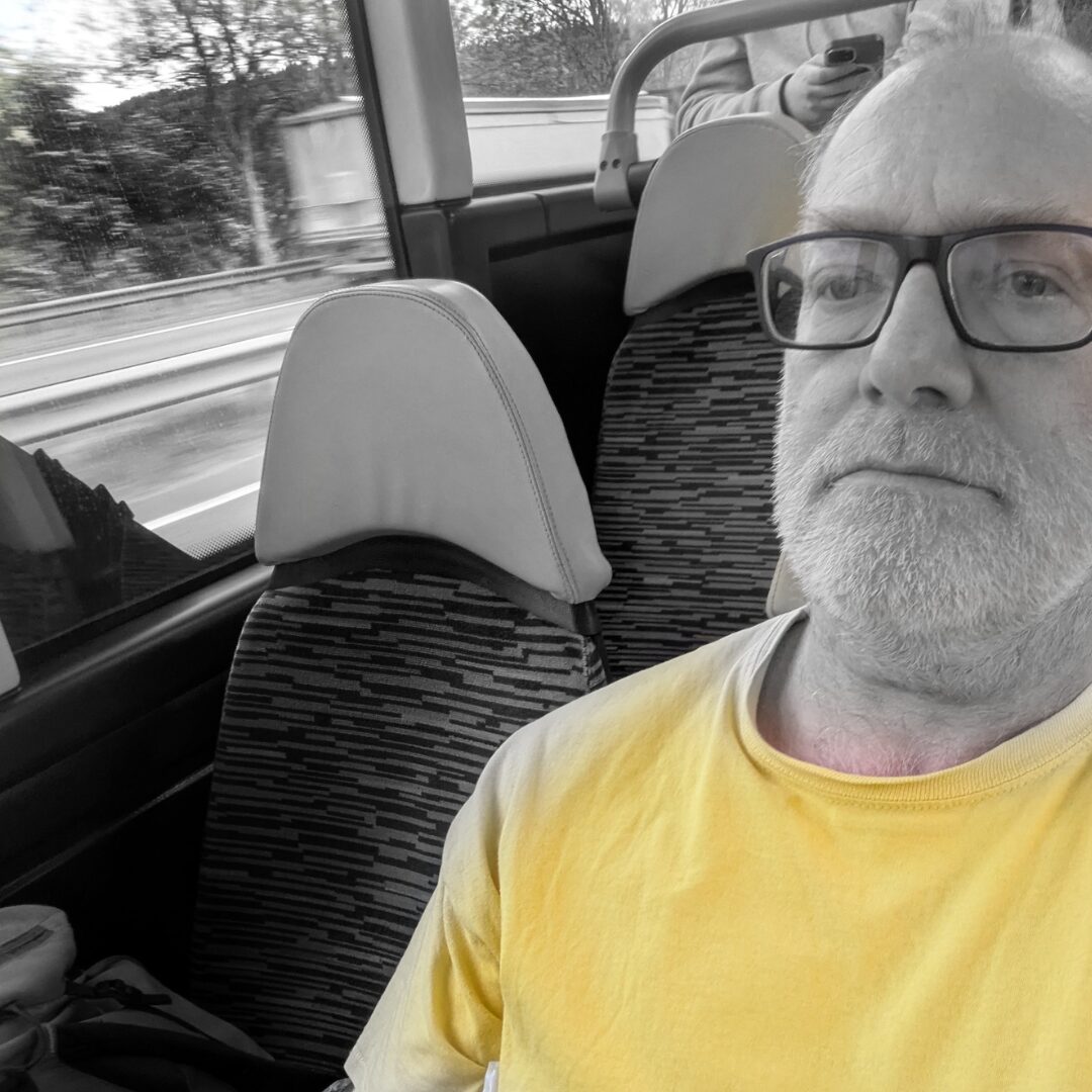 ams colour andrew martyn sugars is dressed in a yellow T shirt. The rest of the image is in black and white. The person wears glasses and is travelling on a bus. They sit in the aisle seat. The seat next to them and behind them is empty. You can see traffic passing through the window.