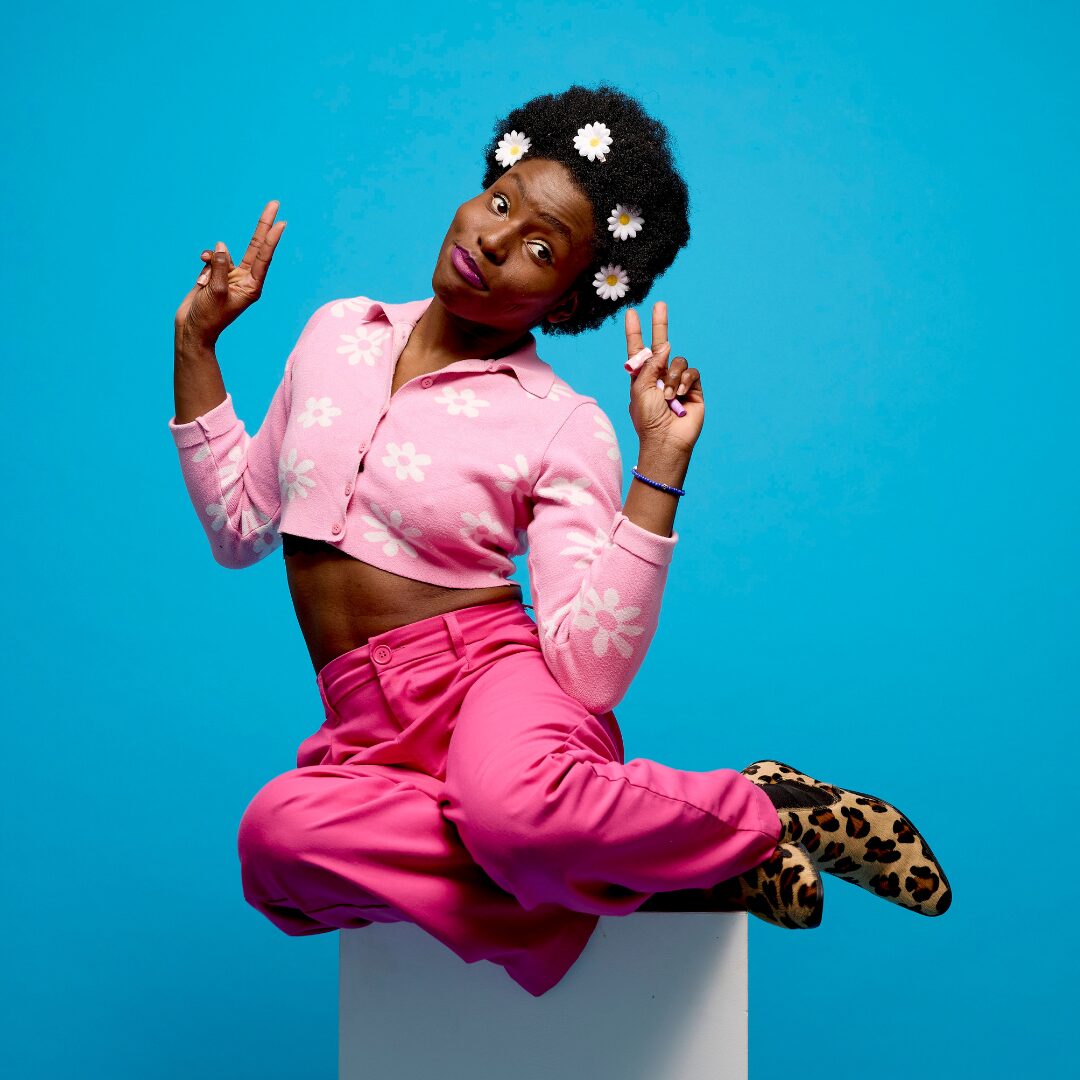 fc colour Farrell Cox is a black person dressed in a pink cropped top with white flowers, pink trousers and animal print shoes. They wear daisies in their short black hair. They perch on top of a white plinth with their legs tucked to the side. The background is bright blue.