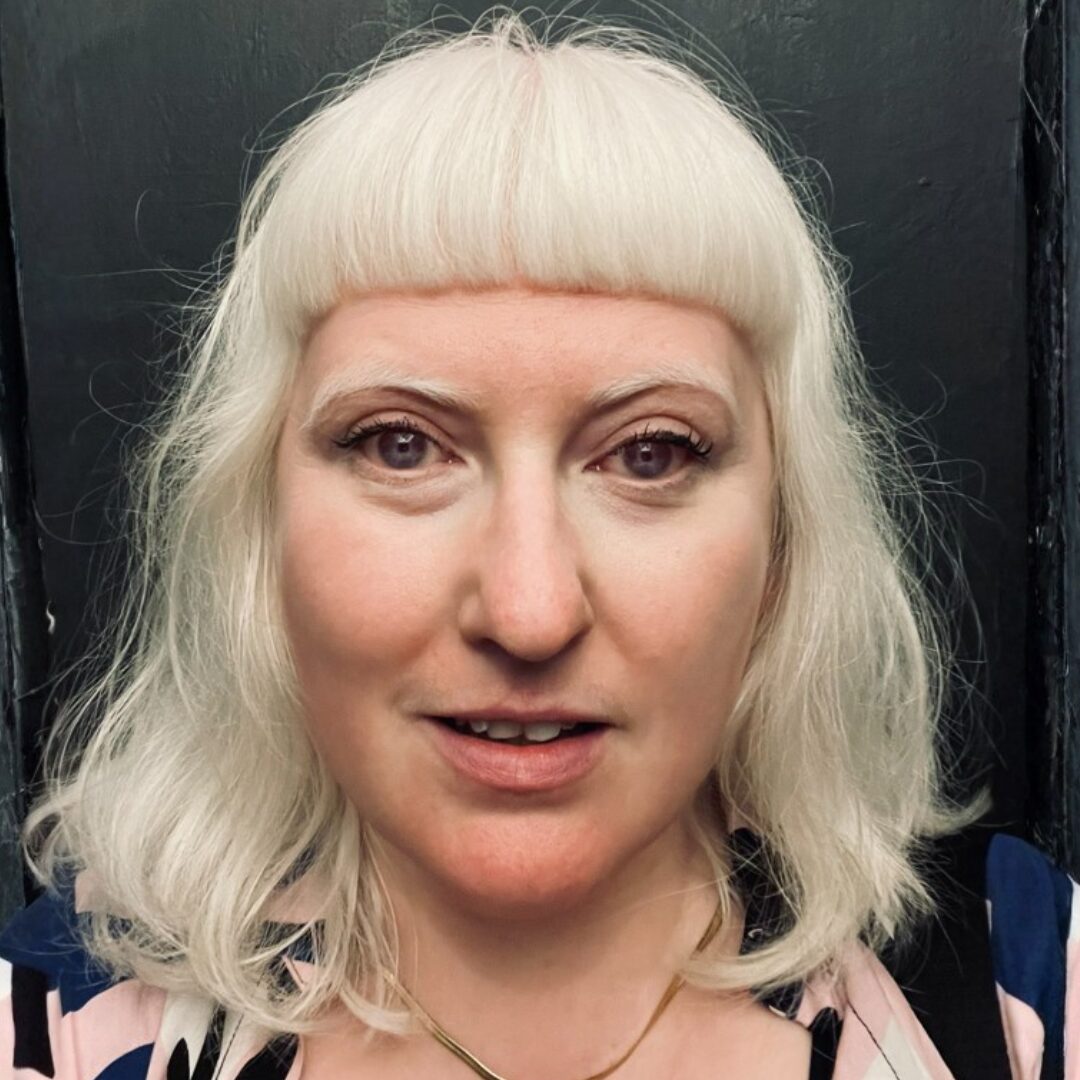 jb colour A headshot of Jo Bannon. They are in front of a black painted background. The have shoulder length blonde hair and a straight, short fringe. They stare straight into the camera.