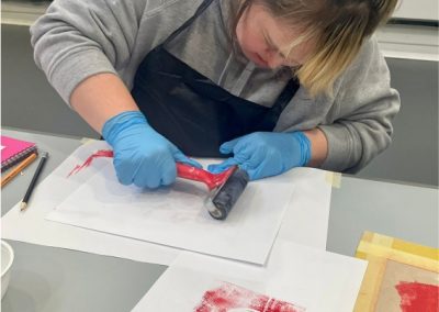 Female artist wearing grey top, black apron and blue gloves printing geometric shapes on to white paper with roller and red ink