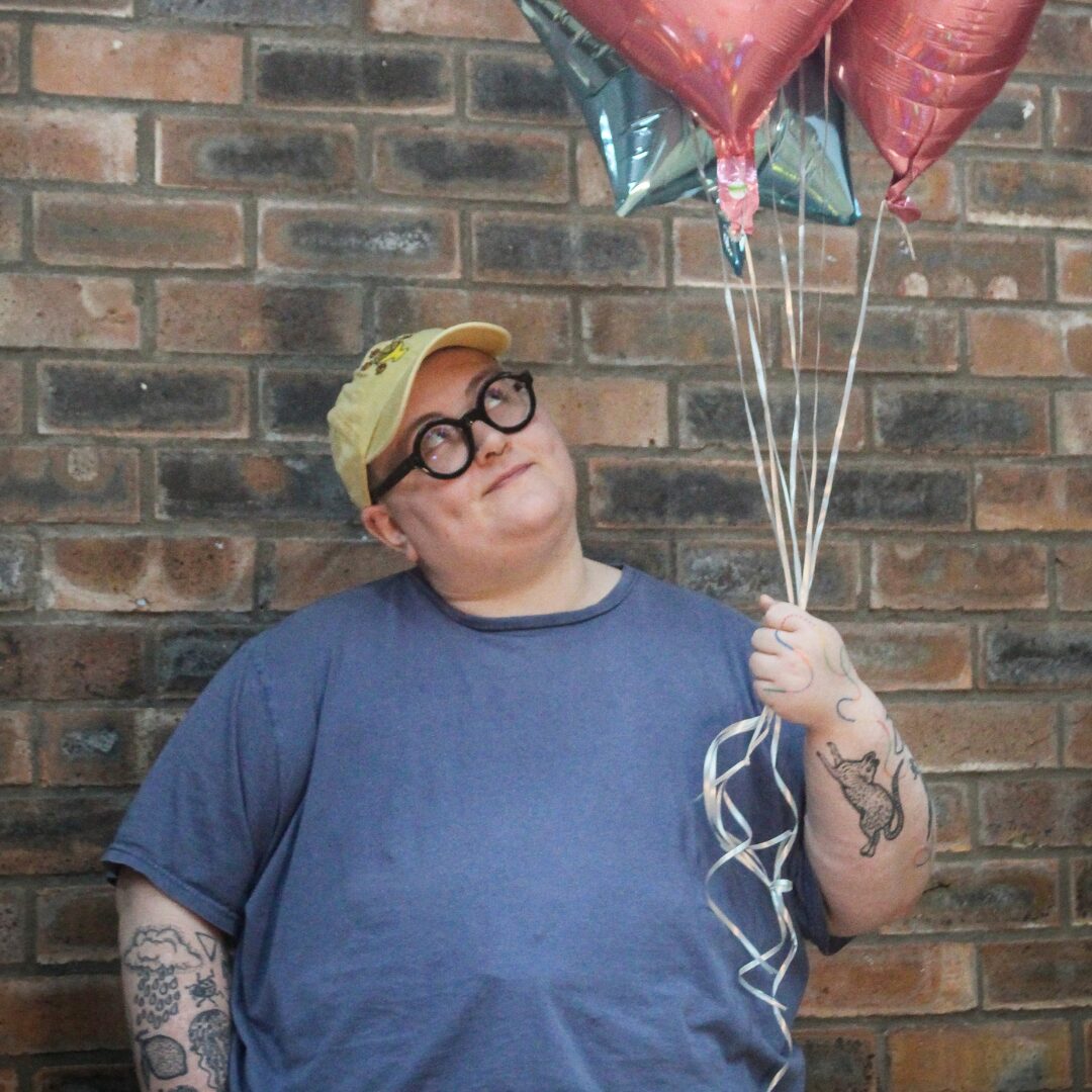 mm colour Maddie Morris stands in front of a brown brick wall. They wear a blue T shirt, yellow baseball cap and black glasses. They hold some pink and blue helium balloons in their hand and they look sideways up to the balloons.